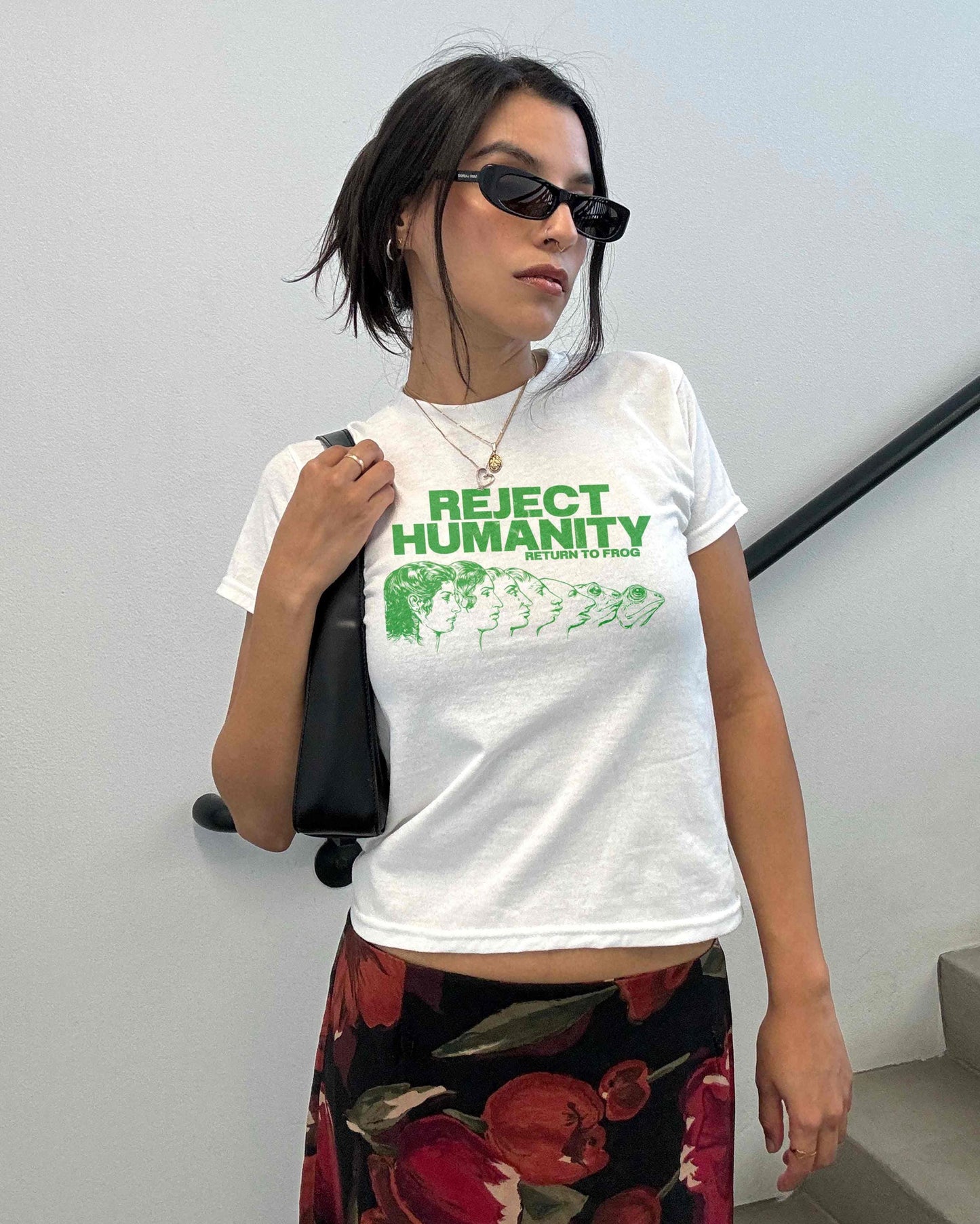 Young woman wearing a white Y2K baby tee, perfect for Gen Z fashion lovers and teens into alt clothing. The shirt features a green graphic print with the text "REJECT HUMANITY RETURN TO FROG" and an illustration showing human faces morphing into frogs. This funny meme shirt taps into internet culture and weirdcore aesthetic. It's a great sarcastic gift for anti-social friends or any frog lover. Ideal casual wear top, this graphic tee suits everyday outfits or alternative style look