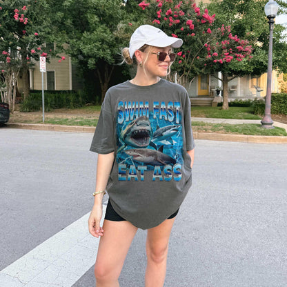 Funny adult humor graphic tee featuring three fierce sharks swimming aggressively with the edgy phrase &quot;Swim Fast Eat Ass.&quot; Vibrant vintage-style design with bold blue letters and ocean wave graphics. Perfect meme culture shirt for young adults, shark lovers, sarcastic friends, or edgy trendsetters. Ideal casual tee for Shark Week, beach trips, parties, or humorous gifts. Provocative yet playful, this shirt makes a bold style statement.