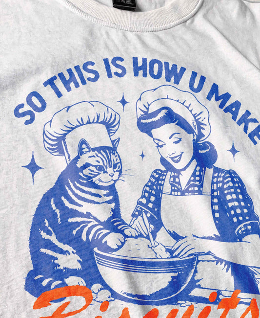 Funny cat t-shirt for men and women featuring a vintage-style illustration of a woman and a cat &quot;making biscuits&quot; together in a mixing bowl. The cat wears a chef hat, humorously mimicking the kneading behavior cats are known for. Retro colors and design give it a nostalgic feel. Great for cat lovers, pet parents, and fans of funny animal shirts. Makes a perfect gift for cat moms, dads, or kitchen humor enthusiasts.