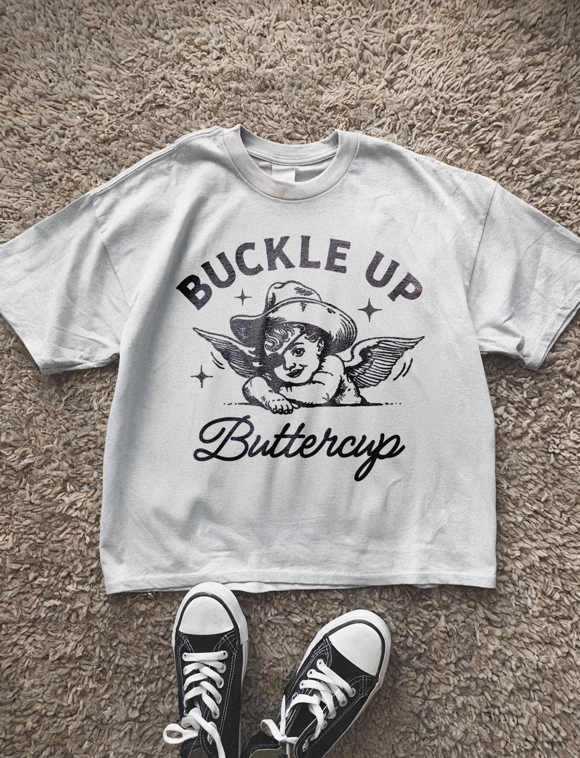 unisex t-shirt featuring a whimsical graphic of an angelic cherub wearing a cowboy hat with the text &quot;Buckle Up Buttercup,&quot; blending playful and vintage western aesthetics for a humorous and charming look.