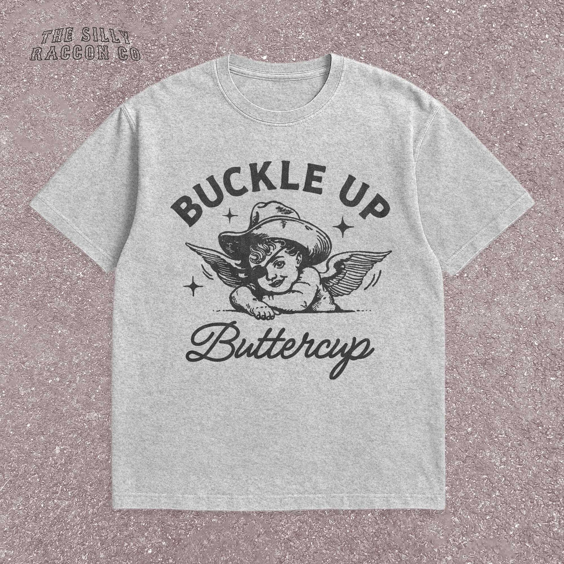 Funny western shirt for women featuring a vintage cowboy angel with wings and the quote &quot;Buckle Up Buttercup&quot; in bold retro typography. The design blends southern sass with angelcore and rodeo aesthetics, perfect for cowgirl fashion or western gifts. Great as a vintage graphic tee with humor and charm for her, ideal for gifting or personal style with a nostalgic Y2K vibe.
