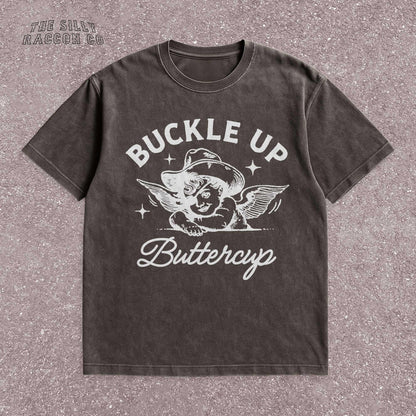 Funny western shirt for women featuring a vintage cowboy angel with wings and the quote &quot;Buckle Up Buttercup&quot; in bold retro typography. The design blends southern sass with angelcore and rodeo aesthetics, perfect for cowgirl fashion or western gifts. Great as a vintage graphic tee with humor and charm for her, ideal for gifting or personal style with a nostalgic Y2K vibe.