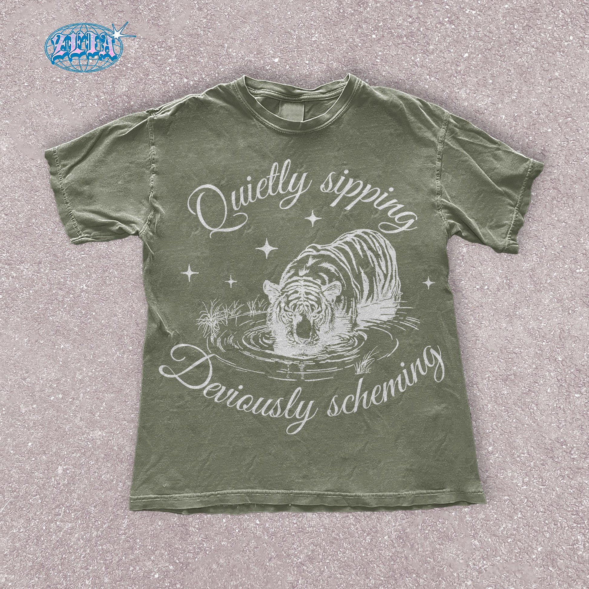 A green moss t-shirt featuring a detailed black graphic of a tiger quietly sipping water with the text Quietly sipping, deviously scheming elegantly curved above and below the tiger.