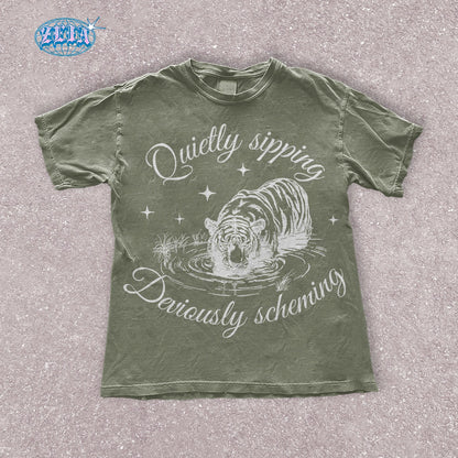 A green moss t-shirt featuring a detailed black graphic of a tiger quietly sipping water with the text Quietly sipping, deviously scheming elegantly curved above and below the tiger.