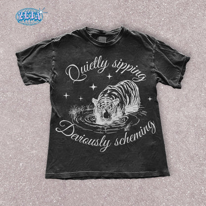 A black t-shirt featuring a detailed black graphic of a tiger quietly sipping water with the text Quietly sipping, deviously scheming elegantly curved above and below the tiger.