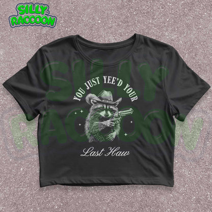 Funny western crop top for women featuring a detailed sketch of a raccoon wearing a cowboy hat and aiming a pistol. The text &quot;You Just Yee&#39;d Your Last Haw&quot; surrounds the raccoon graphic. This humorous graphic tee blends country and meme culture, perfect for young adults and women who love sarcastic animal shirts. Ideal gift for country girls, raccoon lovers, or anyone who enjoys funny southern sayings and western meme apparel.