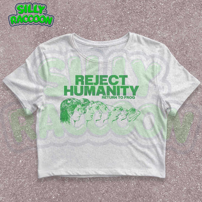 Young woman wearing a white Y2K baby tee, perfect for Gen Z fashion lovers and teens into alt clothing. The shirt features a green graphic print with the text &quot;REJECT HUMANITY RETURN TO FROG&quot; and an illustration showing human faces morphing into frogs. This funny meme shirt taps into internet culture and weirdcore aesthetic. It&#39;s a great sarcastic gift for anti-social friends or any frog lover. Ideal casual wear top, this graphic tee suits everyday outfits or alternative style look