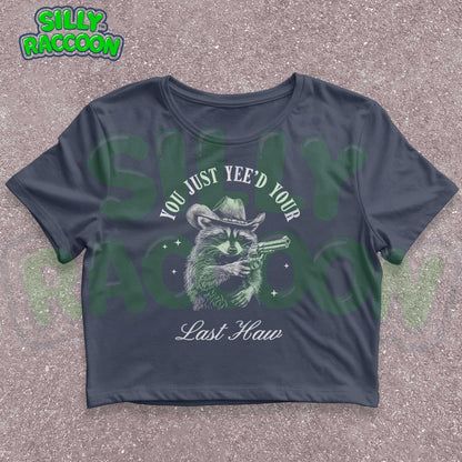 Funny western crop top for women featuring a detailed sketch of a raccoon wearing a cowboy hat and aiming a pistol. The text &quot;You Just Yee&#39;d Your Last Haw&quot; surrounds the raccoon graphic. This humorous graphic tee blends country and meme culture, perfect for young adults and women who love sarcastic animal shirts. Ideal gift for country girls, raccoon lovers, or anyone who enjoys funny southern sayings and western meme apparel.