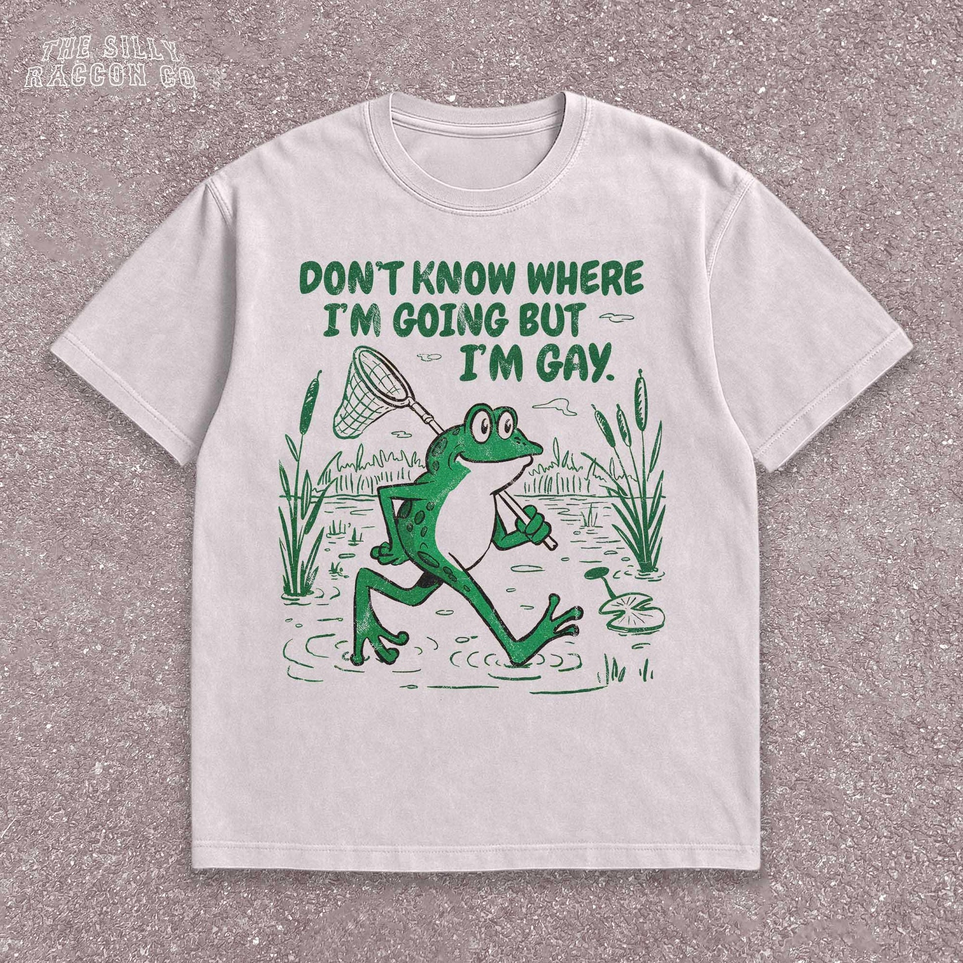 Funny Gay Frog Shirt for men and women featuring a green cartoon frog walking through a marsh carrying a butterfly net over its shoulder. Above the frog, the text reads &quot;DON&#39;T KNOW WHERE I&#39;M GOING BUT I&#39;M GAY&quot; in bold green font. The illustration has a vintage, slightly distressed graphic tee style. This unisex LGBTQ pride t-shirt is a humorous gift for gay friends, perfect for Pride Month or as a coming out gift.