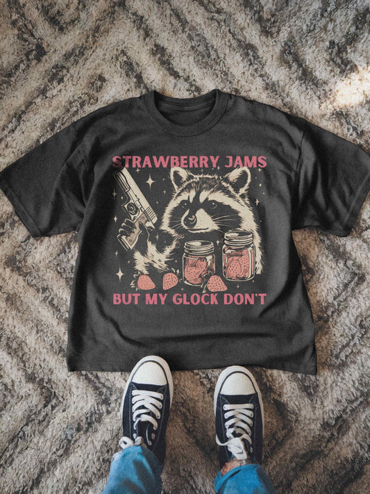 Funny Raccoon Shirt for men and women featuring the meme slogan &quot;Strawberry Jams But My Glock Don&#39;t.&quot; The vintage-style graphic tee shows a cartoon raccoon holding a handgun (Glock) next to two jars of strawberry jam and loose strawberries. The design has a distressed, retro look on a dark grey comfort colors t-shirt. This sarcastic pun shirt makes a great funny gift for him or her, perfect for birthdays, gag gifts, or anyone who loves weirdcore aesthetic, trash panda humor, and me