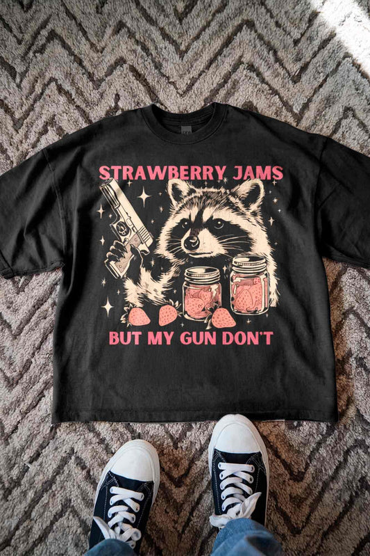 A black t-shirt with a graphic design depicting a raccoon holding a gun and jars of strawberries, accompanied by the text &quot;STRAWBERRY JAMS BUT MY GUN DON&#39;T&quot;. Below the graphic, there is a text that reads &quot;STRAWBERRY JAMS&quot;.